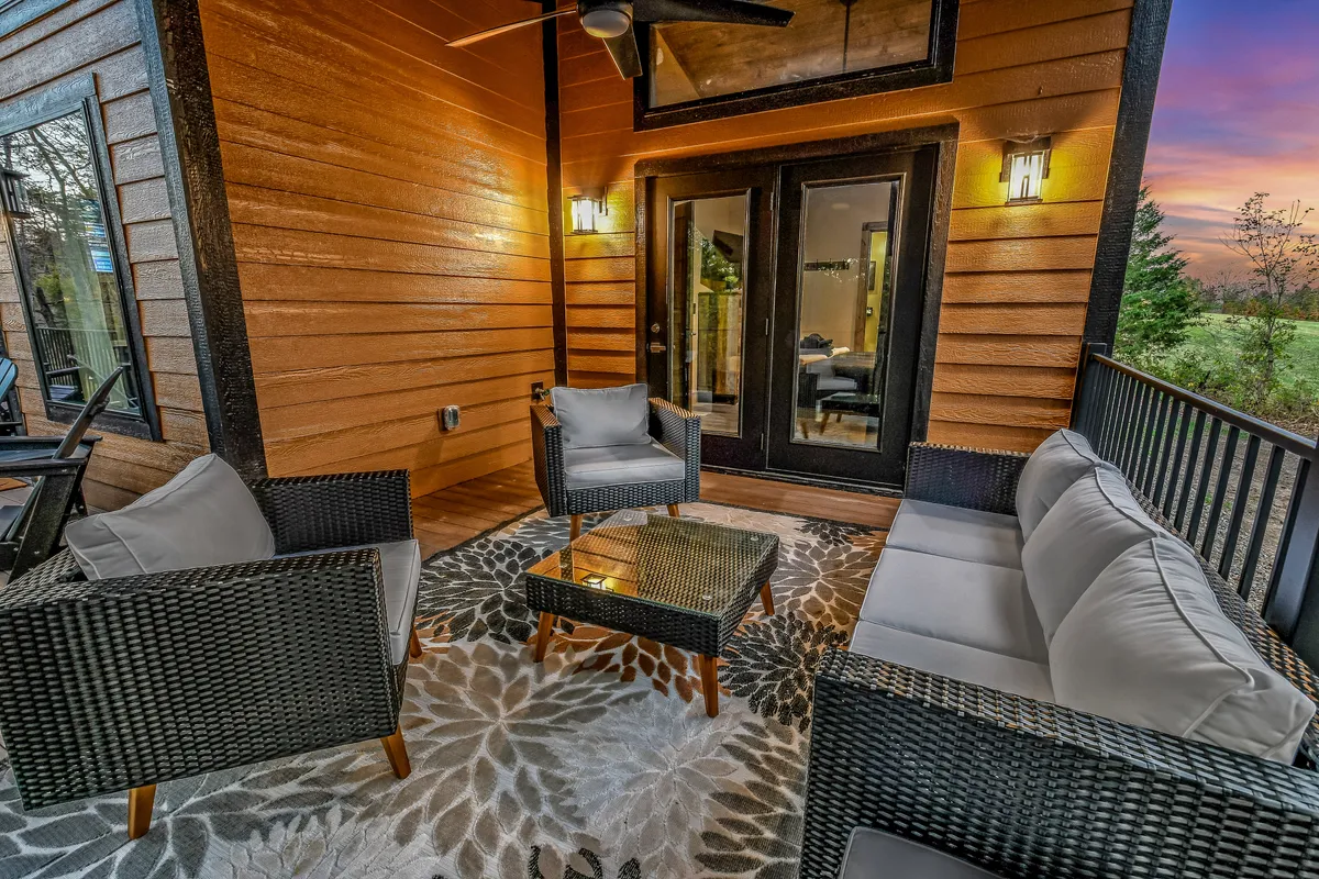 A cozy and stylish outdoor seating arrangement sits atop a decorative area rug, lit by elegant outdoor lighting fixtures. Plush chairs are arranged for easy conversation, with the surrounding environment hinting at tranquil, wooded surroundings. The space is ideal for relaxing evenings under the stars or sharing stories by the firepit.