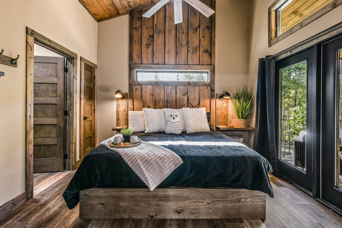 A warm bedroom scene with a welcoming bed, wooden accent wall, large windows allowing in natural light, decorative pillows, and nightstands. The room captures a cozy, inviting vibe ideal for unwinding.