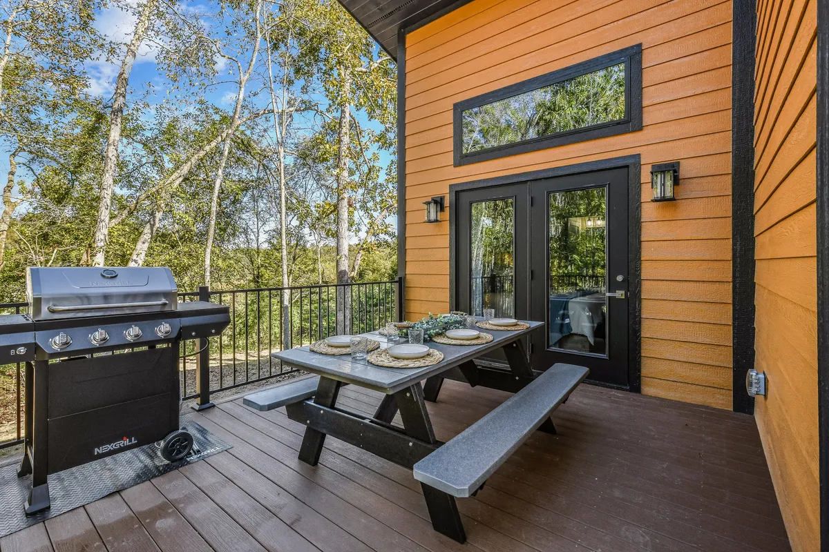 This exterior image showcases a spacious deck with a grill and outdoor dining table set against a backdrop of lush greenery. The setup suggests an inviting area for barbecuing, dining, and socializing while immersed in the natural beauty of the surroundings.