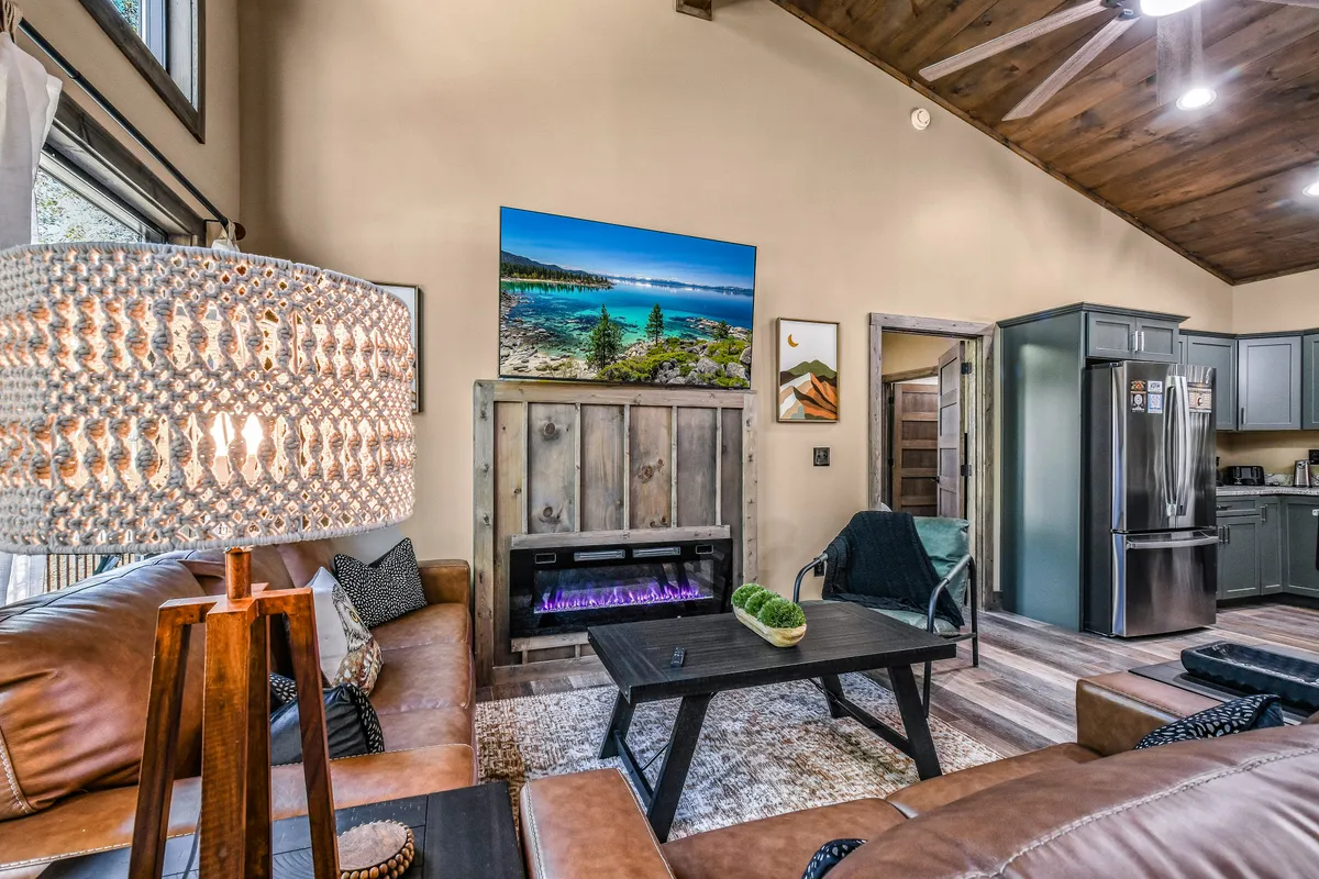 This image features a welcoming interior with a leather sofa, a modern lamp, artistic wall decor, a wood-framed electric fireplace, and a large flat-screen TV. The kitchen area is visible in the background, establishing the open-concept layout. Natural light streams in from large windows, enhancing the inviting atmosphere.