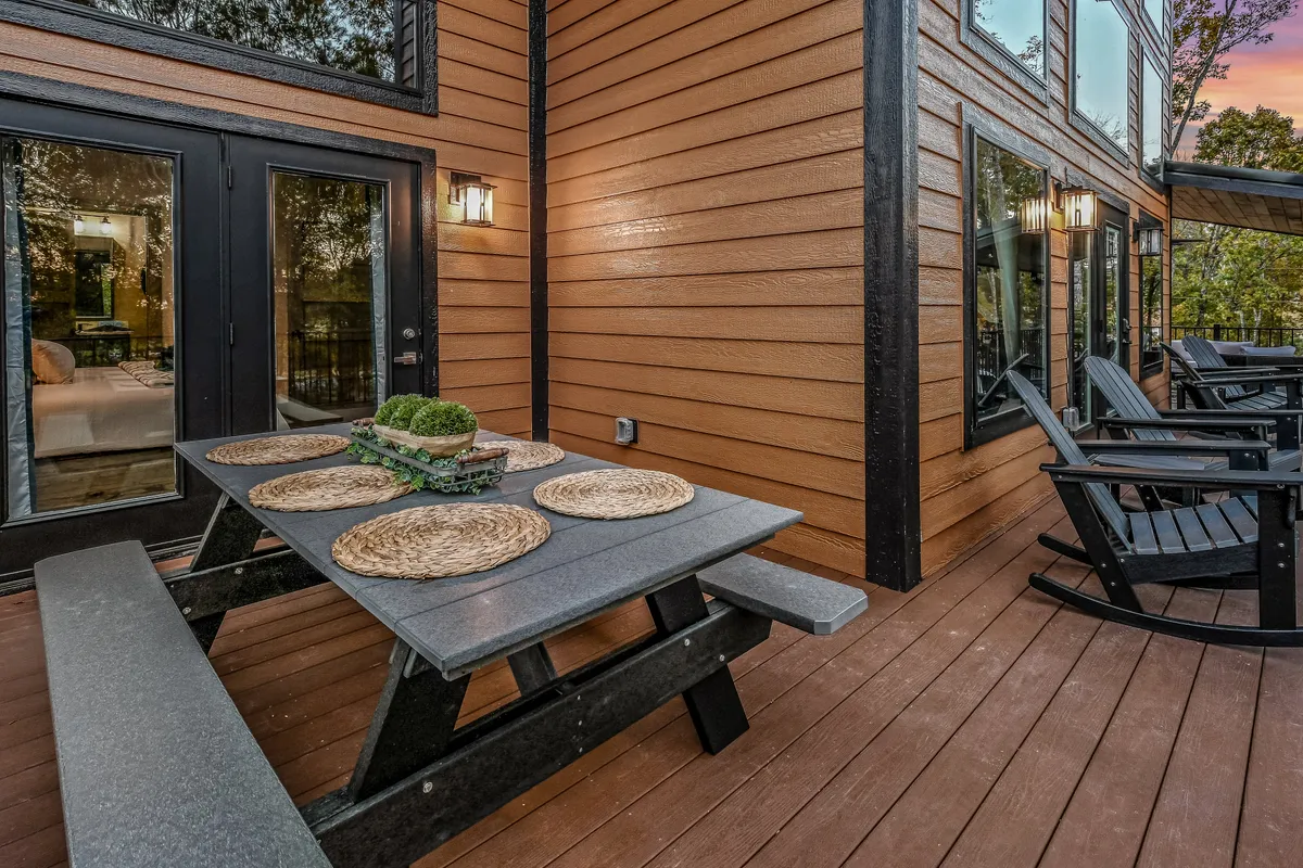An inviting outdoor dining table is set with place settings and black chairs, placed against the backdrop of warm wooden siding. The scene is bathed in soft evening light, suggesting a welcoming environment for outdoor meals and gatherings. The setup encourages shared experiences and conversation in a peaceful natural setting.