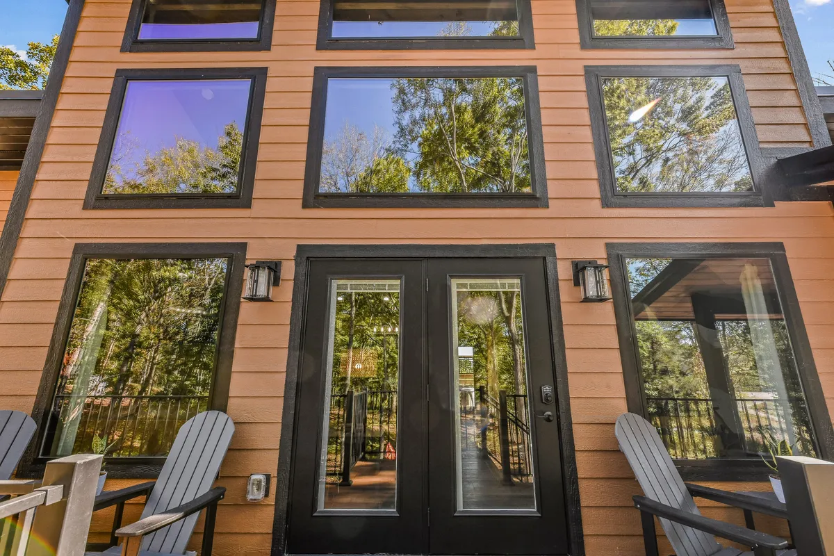 A closer exterior view of the cabin’s large windows, front door, and wooden deck with Adirondack chairs. The image suggests both accessibility and the welcoming nature of the entryway, blending the comfort of the cabin with its natural surroundings.