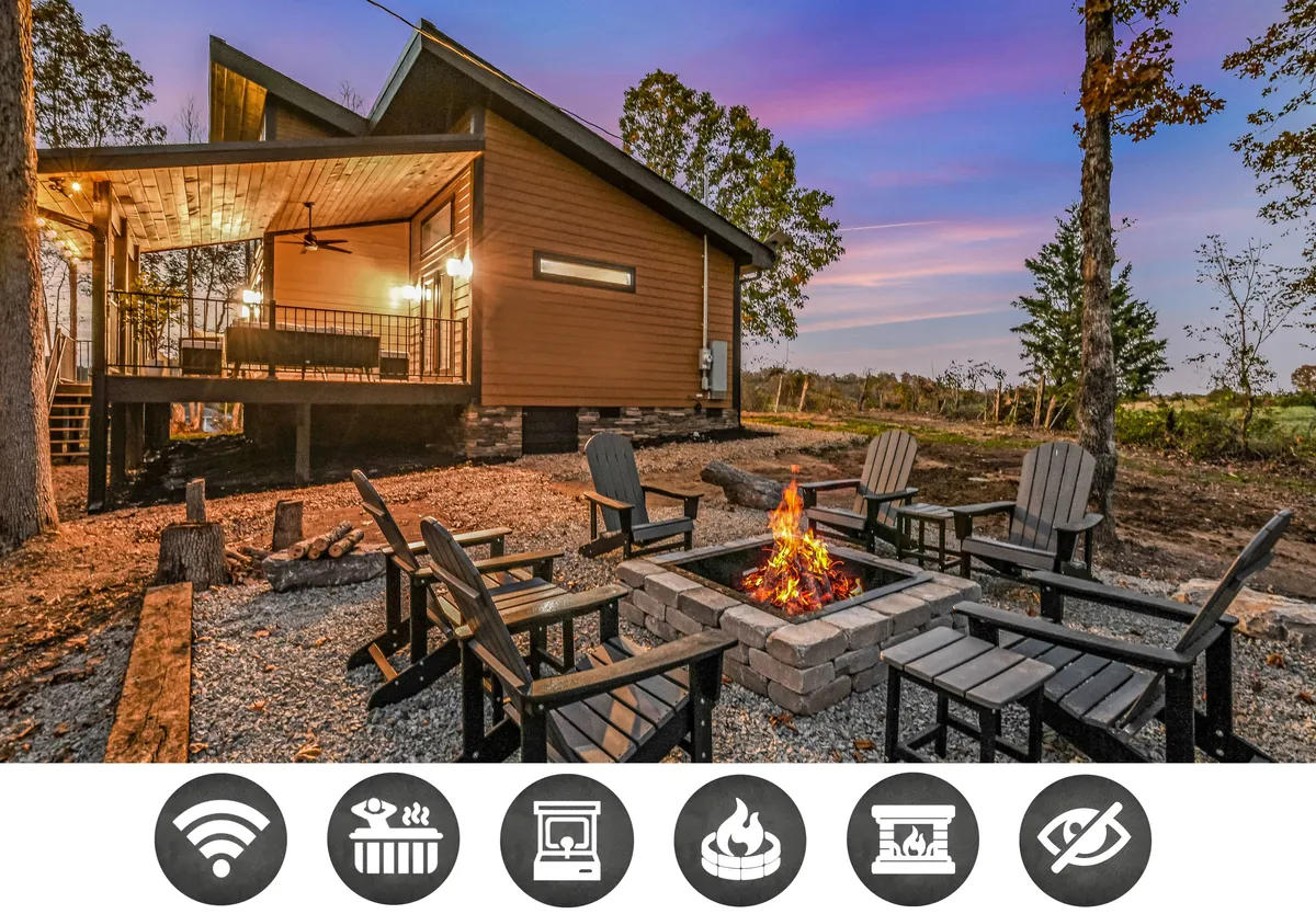 The image showcases the exterior of the Wooded Hideaway cabin, with a glowing fire pit surrounded by comfortable chairs on a spacious deck nestled among the trees. The inviting setup promises a perfect spot for guests to gather, share stories, and enjoy the tranquil forest setting under the stars.