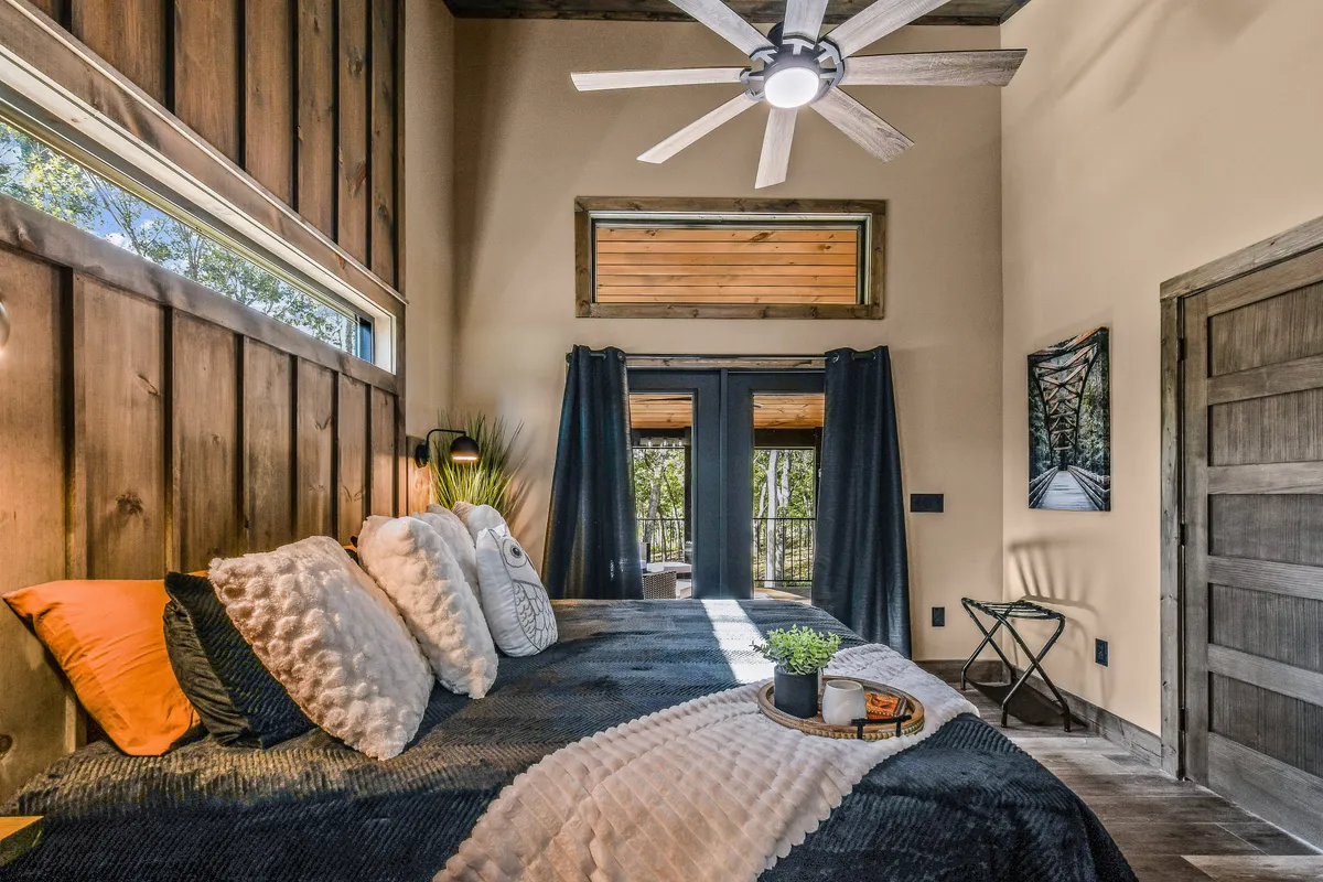 A tranquil king bedroom featuring a plush bed with decorative pillows, large windows framed by dark curtains, a wooden ceiling, and tasteful wall art. The room exudes comfort, with gentle natural light and serene views visible outside.