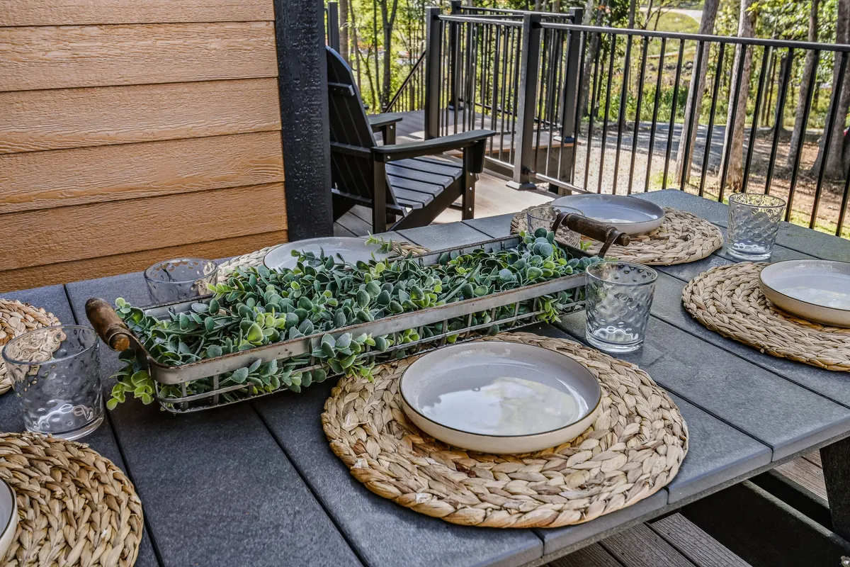 A sunlit deck features a dining table with a greenery centerpiece, woven placemats, and clear glasses, all set for a casual meal. The natural wood and surrounding foliage create a seamless transition between the home and the outdoors, inviting guests to enjoy group meals in a tranquil setting.