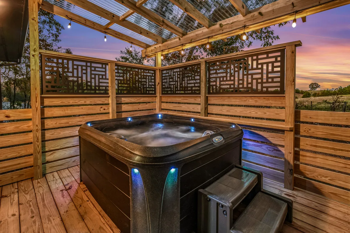 A covered hot tub sits on a wooden deck, partially screened for privacy, with a tranquil landscape of trees in the background. Warm ambient lighting and comfortable surroundings create an inviting spot for relaxation, ideal for unwinding after a day of outdoor adventure.