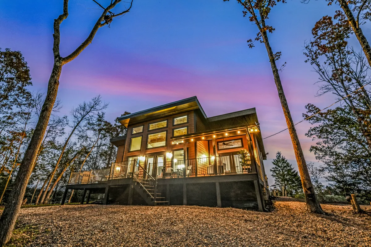 This exterior shot highlights the modern architecture of Wooded Hideaway, showcasing large windows, a well-lit entryway, and the spacious, level gravel driveway surrounded by trees. The scene conveys a welcoming, tranquil arrival experience.