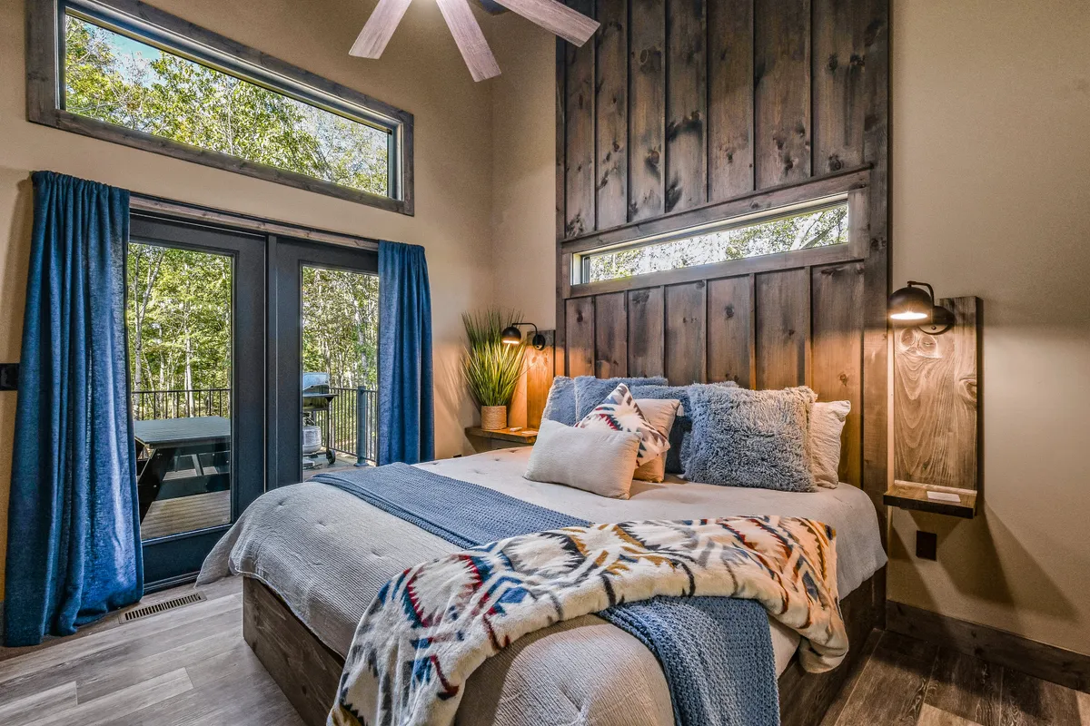 A cozy bed with decorative pillows is placed against a wooden accent wall, with large windows that open to beautiful outdoor nature views. The setup is warm and inviting, perfect for restful sleep or snuggling up with a view of the trees.