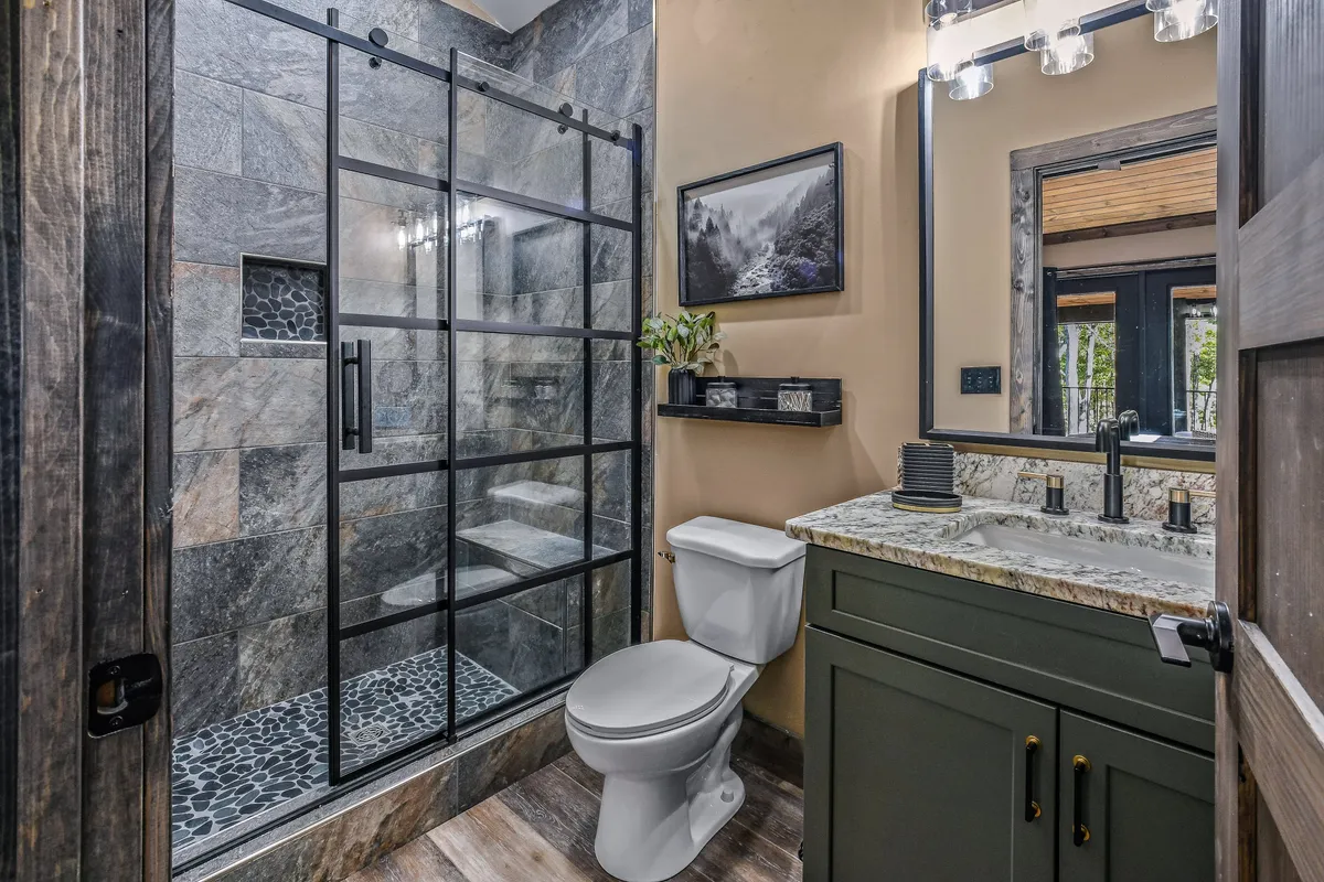A modern, serene ensuite bathroom with a glass shower enclosure, granite countertop, framed artwork, and a potted plant. The space is bright and calming, with high-end finishes.