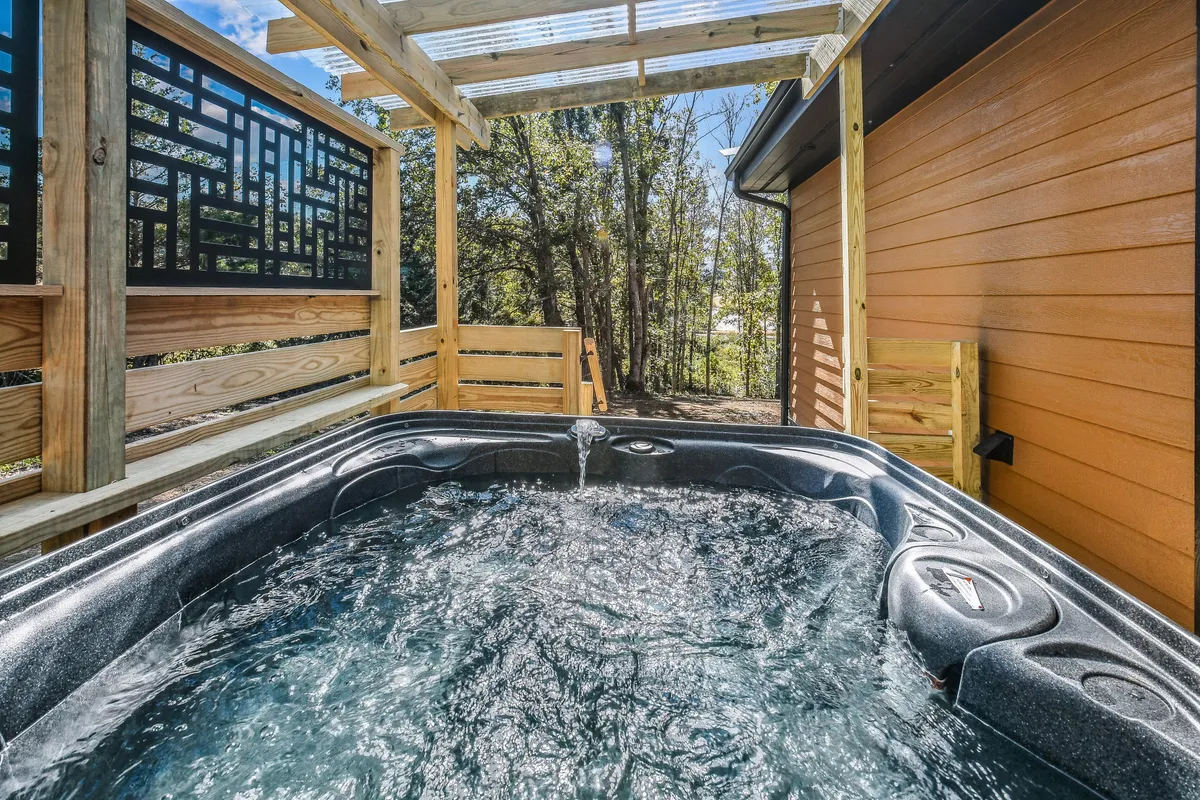 A private hot tub is nestled beneath a wooden structure, enveloped by tall trees and lush foliage. The peaceful setting offers a tranquil space for guests to unwind and recover after a day of exploration.