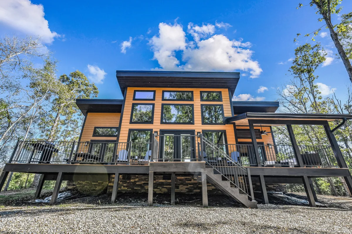 The exterior of the property showcases large glass windows, a spacious wooden deck, and modern architecture nestled among mature trees. The inviting outdoor seating area and expansive deck suggest a space designed for group gatherings with easy access to nature.