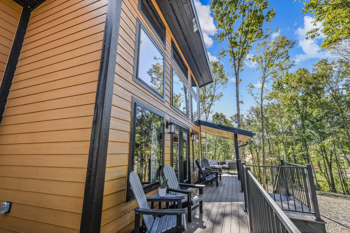 A wooden deck wraps around the house, bordered by large windows and surrounded by tall, leafy trees. The scene captures late afternoon light streaming through the forest, with comfortable seating areas visible, inviting relaxation while connecting seamlessly to the natural surroundings.
