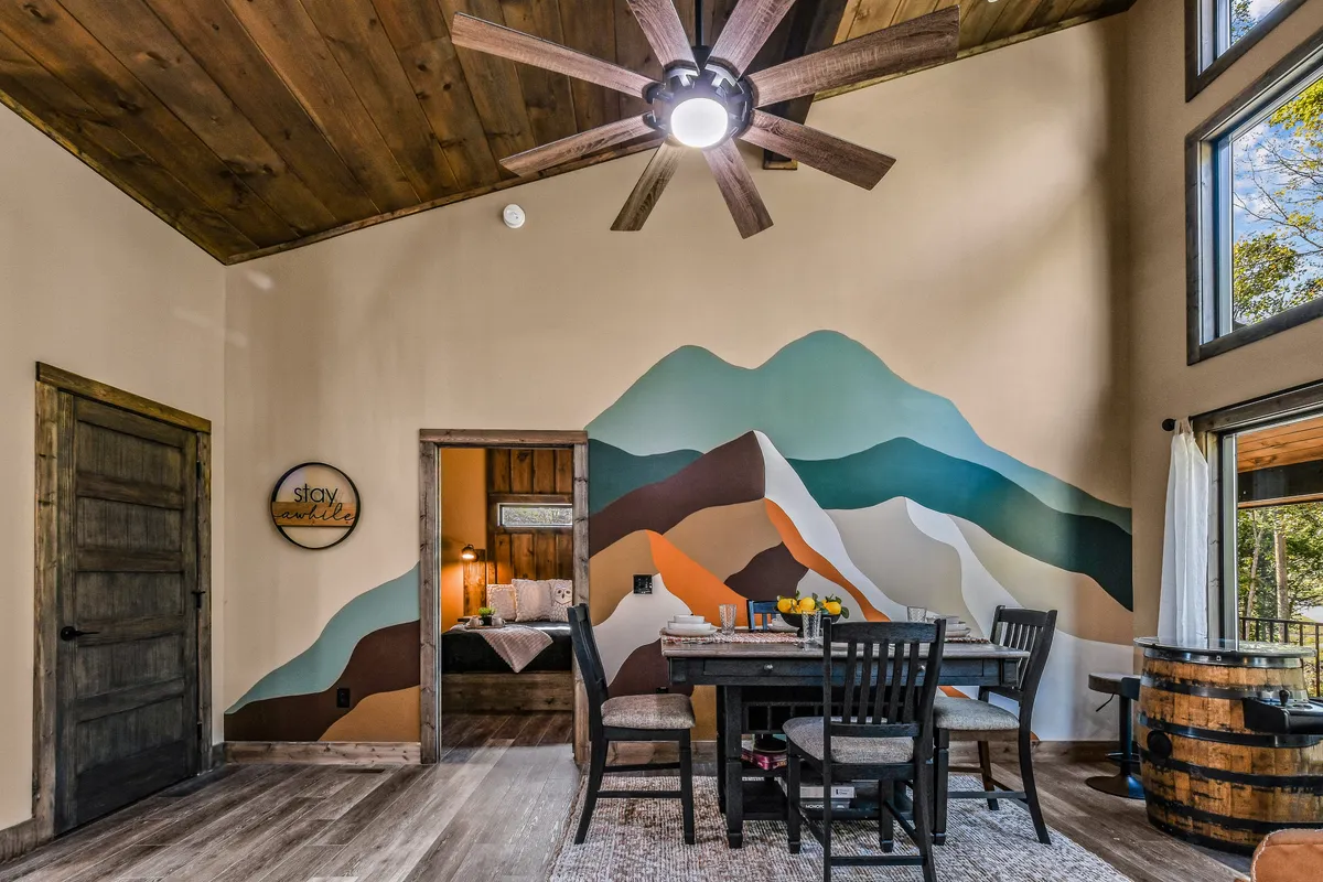 A spacious dining area is featured with a black dining table and chairs, set against a striking hand-painted colorful mountain mural. Large windows behind the table allow natural light to flood in, connecting the indoors to the surrounding forest views.